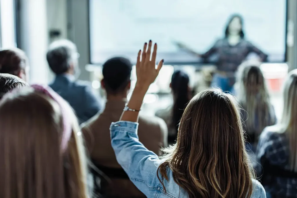 En gruppe mennesker i et klasserom rekker opp hendene som svar på en presentasjon.
