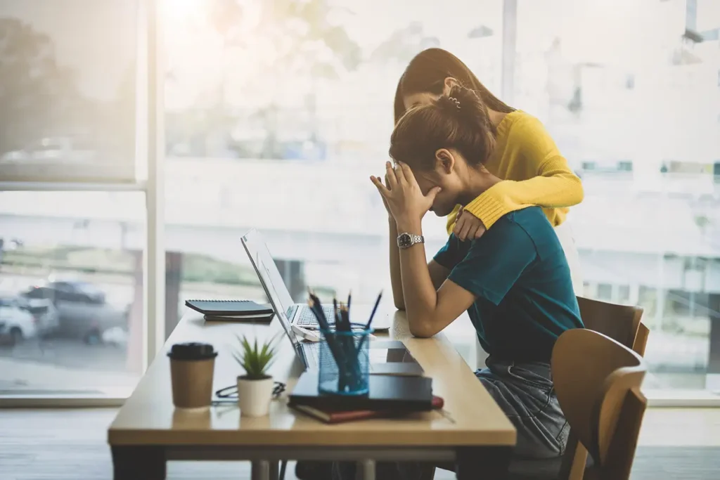 En kvinne i en gul genser trøster en fortvilet kollega som sitter ved et skrivebord på et lyst kontor, omgitt av bærbare datamaskiner og skrivesaker, og diskuterer stress og arbeidsrelatert fravær.