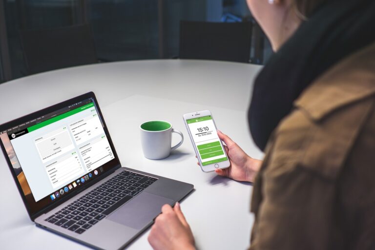 Person som bruker en smarttelefon og bærbar datamaskin ved et bord med et kaffekrus. Begge enhetene viser brukergrensesnitt med grønt tema, noe som reflekterer en forpliktelse til bærekraftig praksis og fremme tillit i arbeidslivet.