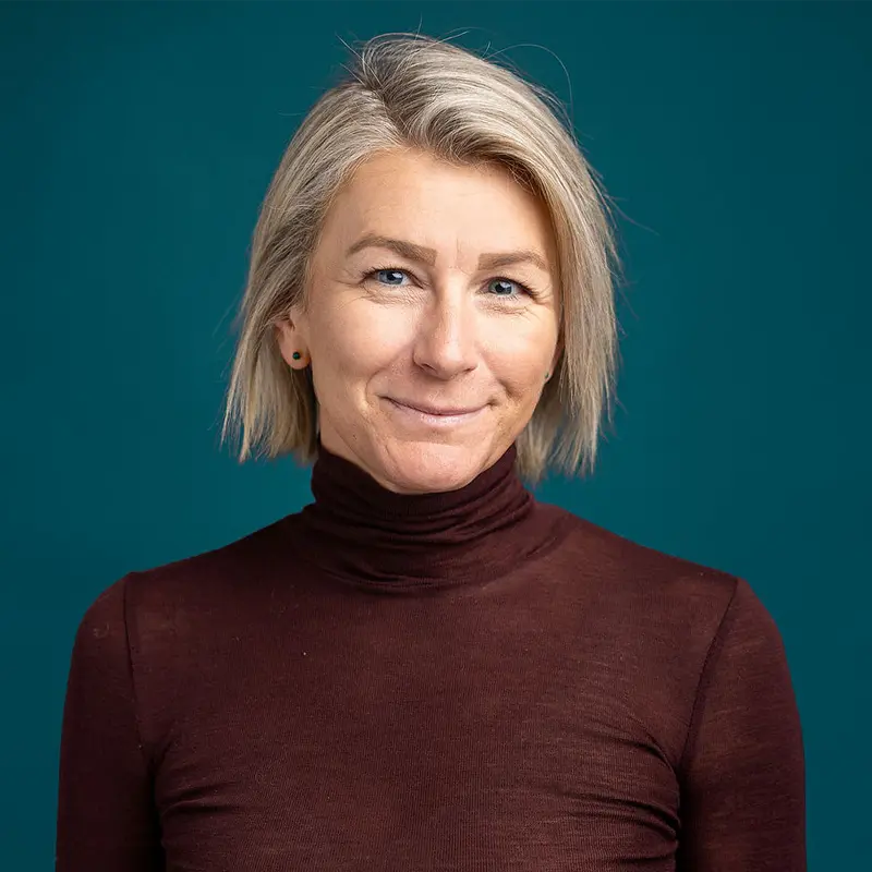 En person med kort blondt hår, iført mørk turtleneck, smiler lett mot en blågrønn bakgrunn, og legemliggjør den profesjonelle essensen til Ansatte Fossekall.
