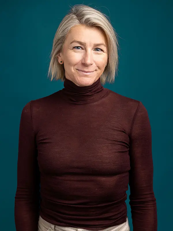 Veslemøy Eineteig Wedum, med kort blondt hår, smiler lett mens han tar på seg en mørkebrun turtleneck mot en solid blågrønn bakgrunn.