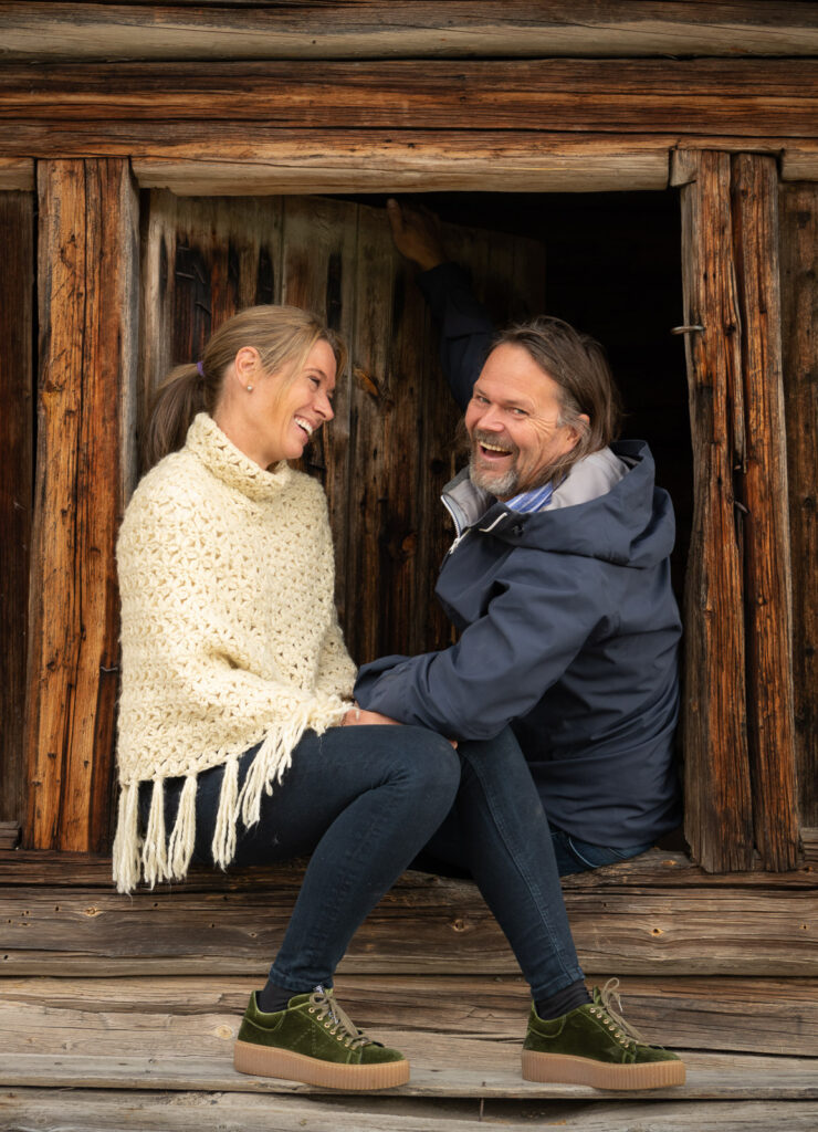 To voksne sitter i døråpningen til en rustikk trehytte på Skåbu Fjellhotell, vendt mot hverandre og smiler. Kvinnen har på seg en kremfarget strikket poncho, og mannen har på seg en blå jakke og jeans.