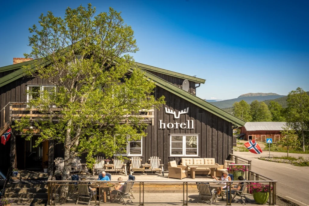 Et rustikt trehotell med et «hotell»-skilt, uteservering og norske flagg, mot en klar blå himmel og fjell i bakgrunnen – velkommen til Skåbu Fjellhotell.