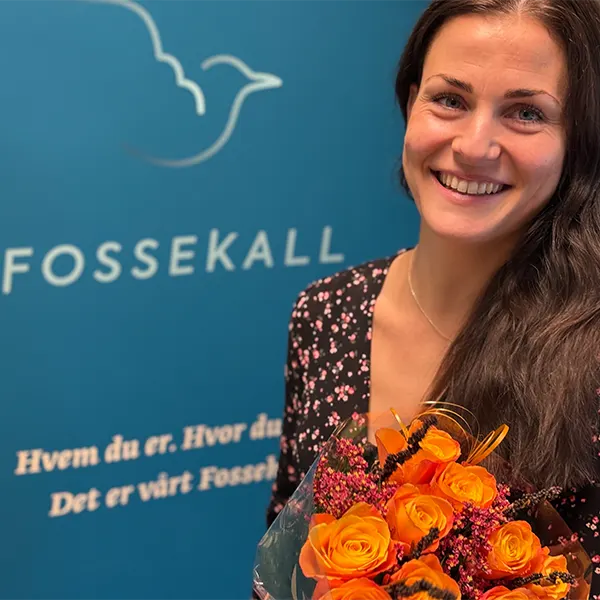 En kvinne med langt, mørkt hår, iført en svart, blomstret topp, smiler mens hun holder en bukett oransje roser foran en blå vegg med teksten "FOSSEKALL", som representerer Ansatte Fossekall.