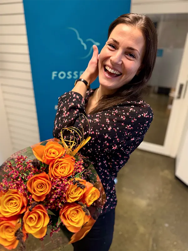 En kvinne smiler med en bukett oransje roser i hånden, mens hun står innendørs ved et blått skilt og en åpen dør på Kjetil Veen-arrangementet.