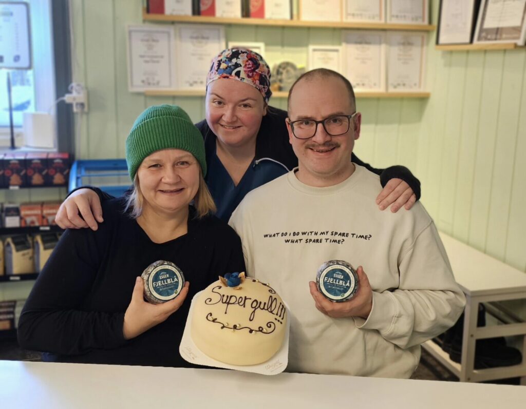 Tre personer poserer innendørs, to holder krukker og én holder en kake merket "Supergull OST". På veggen bak dem henger det sertifikater og utmerkelser.