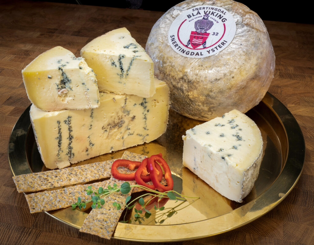 Et hjul og skiver av blåmuggostost er anrettet på et gullbrett med knekkebrød, skiver av rød paprika og mikrogrønt på et underlag av tre. Osten er merket "Snertingdal Ysteri".