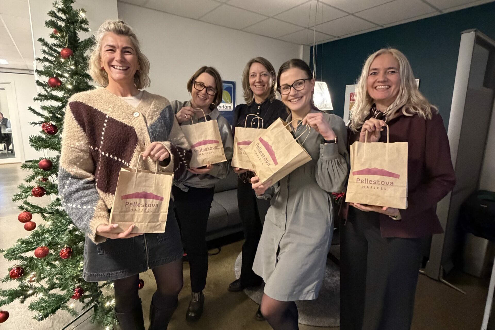 Fem kvinner står innendørs ved et pyntet juletre, holder hver sin papirpose og smiler til kameradette gjorde fossekall i 2025.