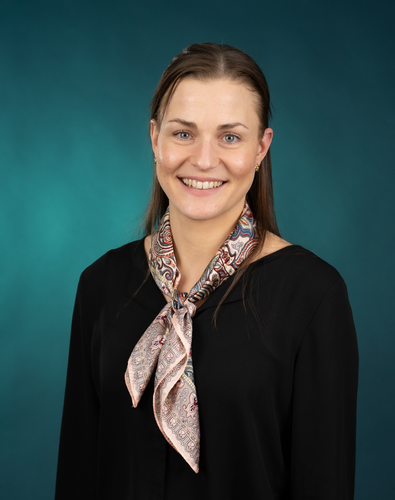 Livia Sauter, en kvinne med rett, brunt hår, iført svart topp og mønstret skjerf, smiler til kameraet mot en blågrønn studiobakgrunn.