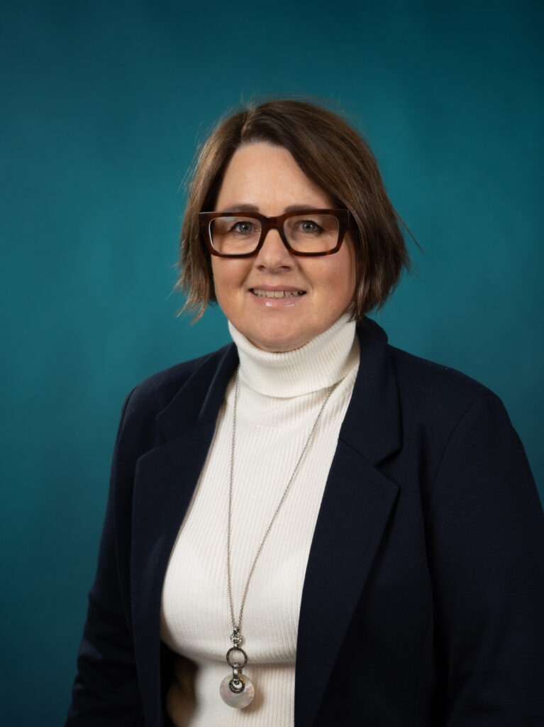 En kvinne med kort, brunt hår, briller, hvit rullekrage, marineblå blazer og langt halskjede poserer mot en ensfarget blågrønn bakgrunn - et eksempel på den profesjonelle stilen man ser hos Ansatte Fossekall.