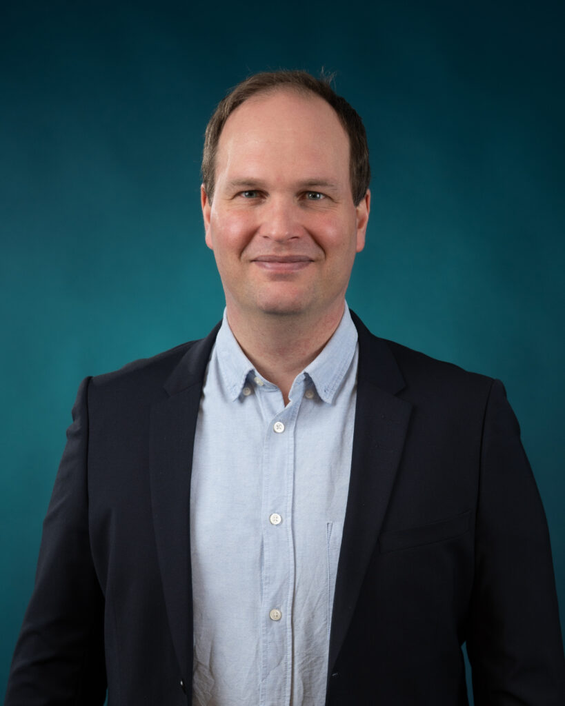 En mann i marineblå blazer og lyseblå skjorte står foran en blågrønn bakgrunn og ser inn i kameraet med et nøytralt uttrykk - et ideelt portrett for Ansatte Fossekall.