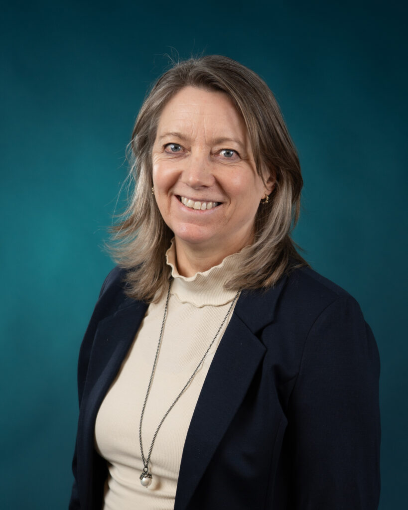 En kvinne med skulderlangt, lysebrunt hår, iført kremfarget turtleneck, mørk blazer og halskjede, smiler til kameraet mot en blågrønn bakgrunn - et innbydende ansikt blant Ansatte Fossekall.