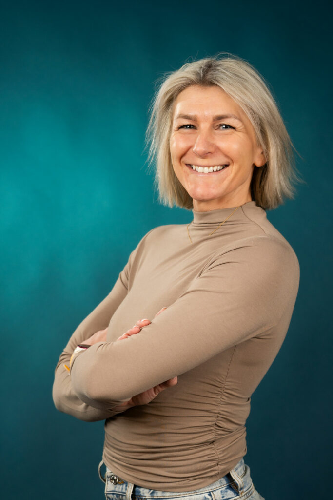 Veslemøy Eineteig Wedum, en kvinne med blondt hår, iført beige langermet topp og lyse jeans, står mot en blågrønn bakgrunn med armene i kors og smiler til kameraet.