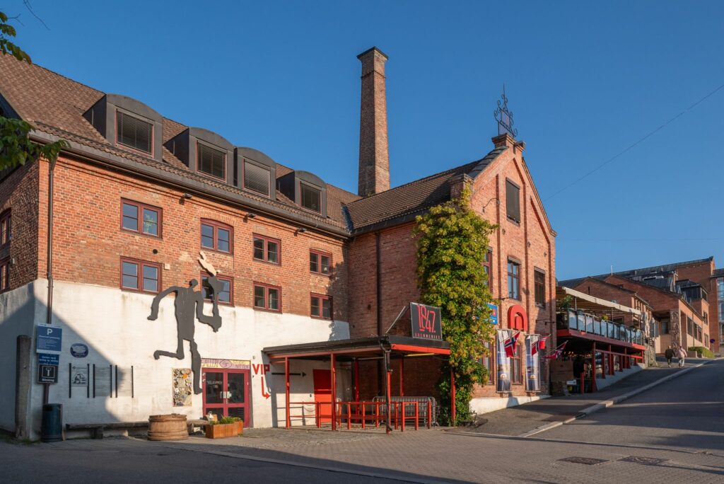 En murbygning med store vinduer, en høy skorstein og et svart silhuettmaleri på veggen; røde dører og en skyggefull uteservering - perfekt for møter hvis du trenger å leie inn operative ressurser på en solrik dag.